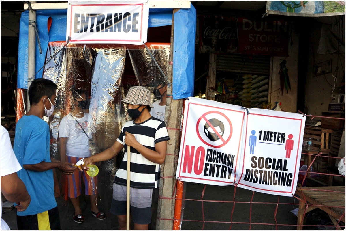 Antipolo City, Philippines - March 31, 2020: A volunteer stands guard at the entrance of a public market to spray alcohol to hands and ensure the use of face mask during the COVID-19. Image Credit: junpinzon / Shutterstock