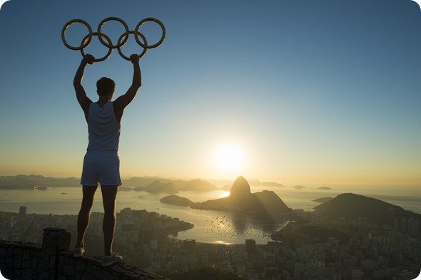 RIO DE JANEIRO, BRAZIL - MARCH 05, 2015 Illustrative editorial of man standing in silhouette holding Olympic rings above city skyline view of Sugarloaf Mountain and Guanabara Bay at sunrise-lazyllama