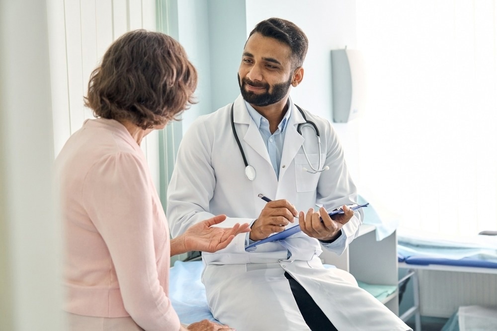 Professional physician wearing white coat talking to mature woman signing medical paper at appointment visit in clinic. Image Credit: Ground Picture/Shutterstock.com