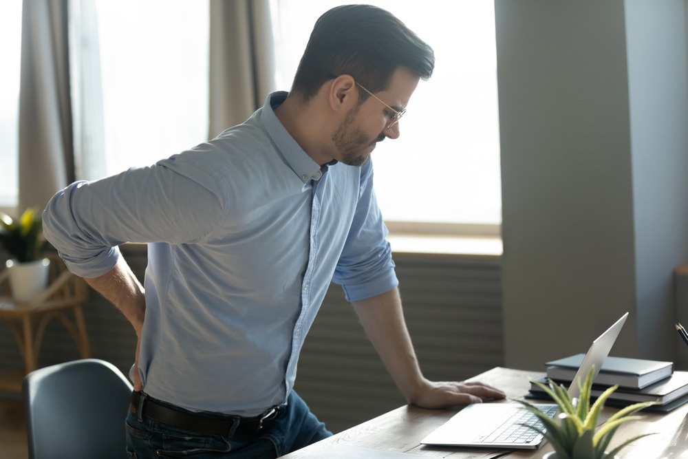 Businessman touches lower back feeling pain sudden ache while get up out of office chair, muscular spasm, strain caused by sedentary lifestyle, prolonged inactivity, sit in incorrect posture concept. Image Credit: fizkes/Shutterstock.com
